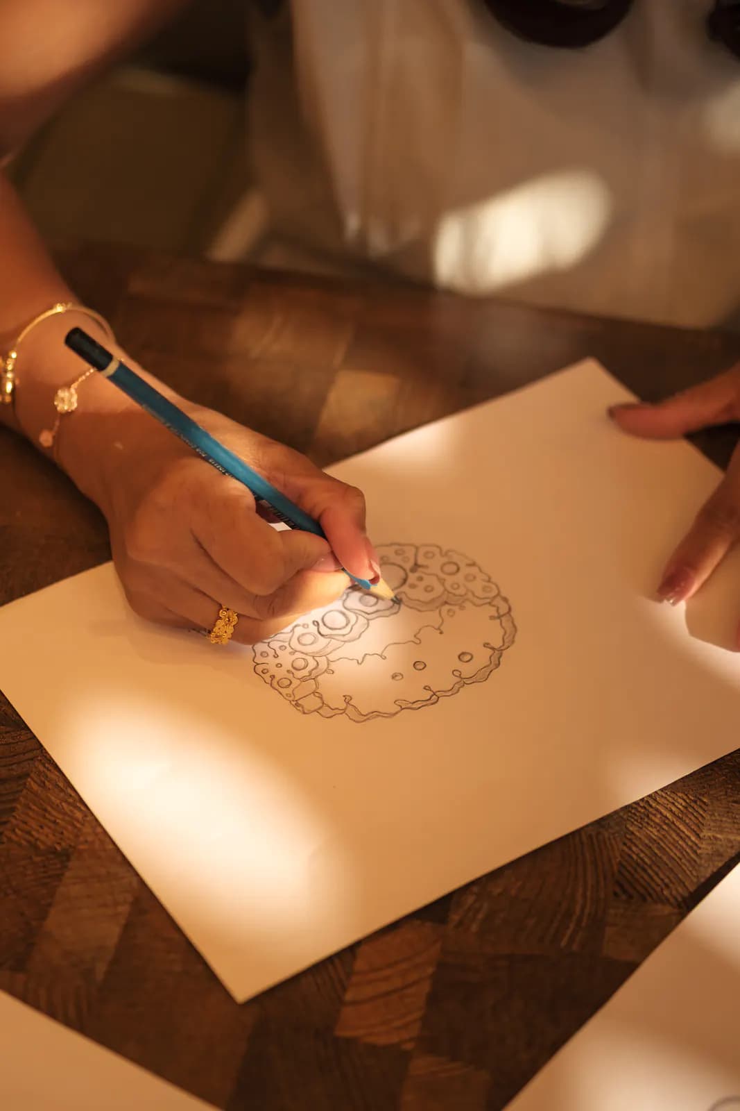Hand wearing gold jewelry sketching a floral design on paper
