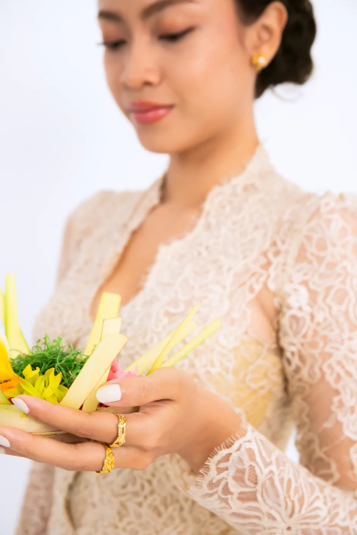 Model in a lace kebaya smiling down at a canang offering, wearing gold rings