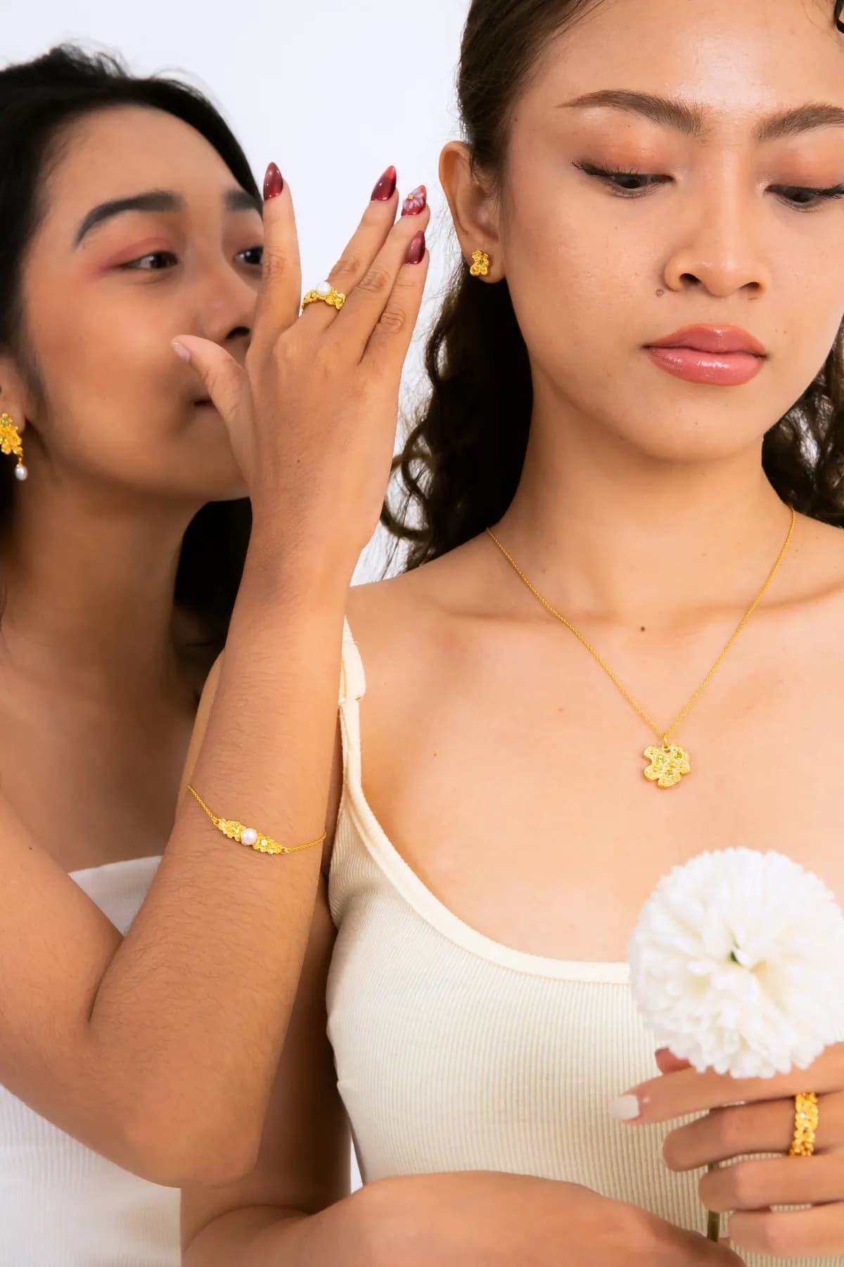 Two models whispering, one wearing a gold flower pendant and holding a white flower