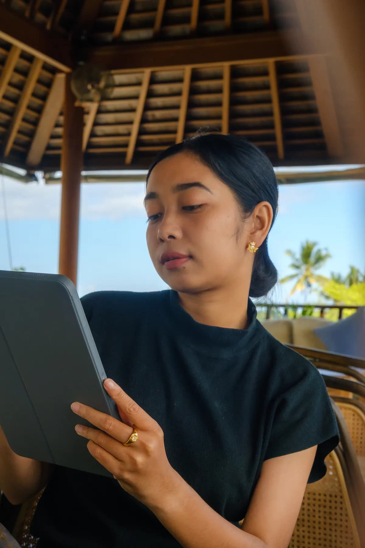 Woman reading a tablet under a thatched bale, wearing gold earrings and a ring