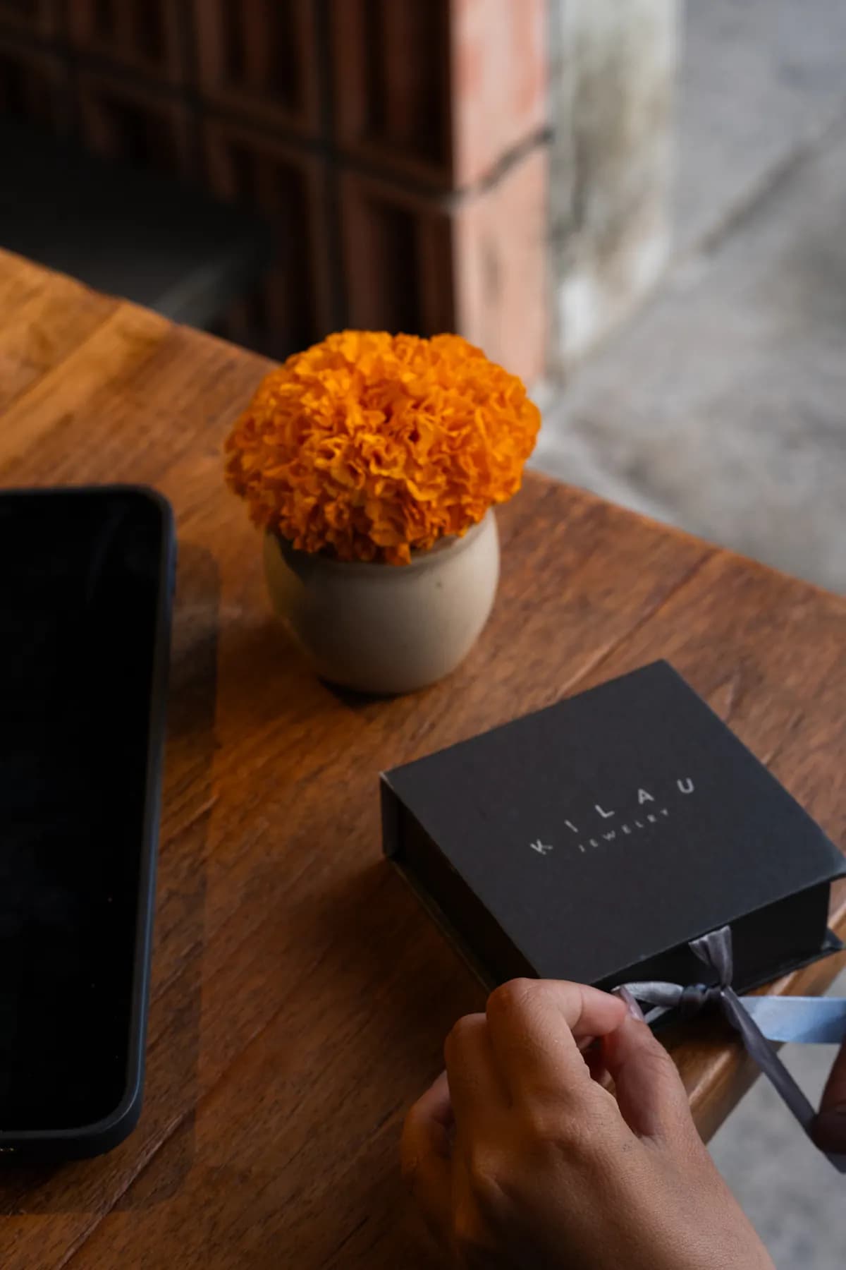 Hands opening a black KILAU gift box on a wooden table beside marigolds